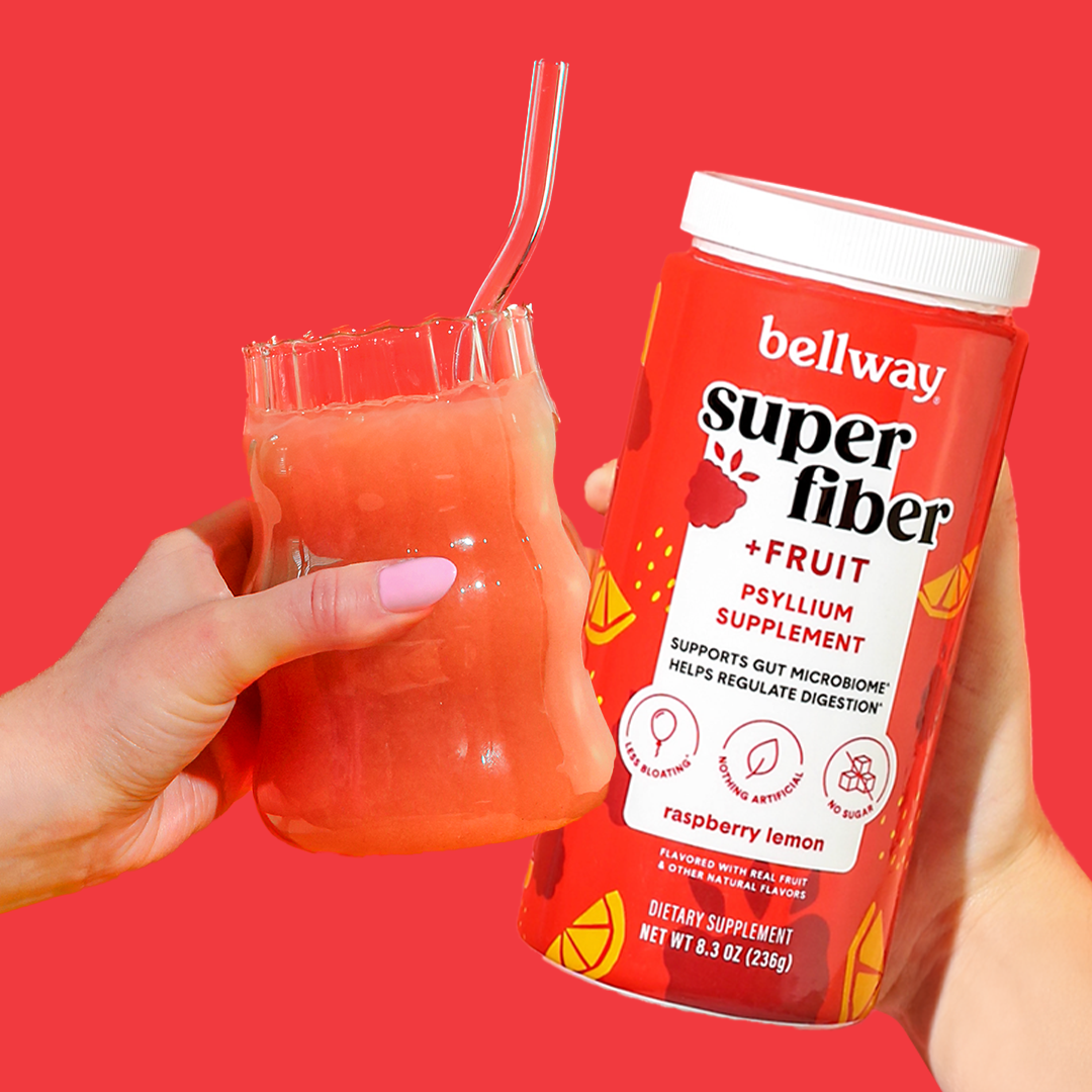 A hand holding a Bellway Super Fiber + Fruit raspberry lemon tub meets another hand holding the supplement drink in a clear glass on a red background