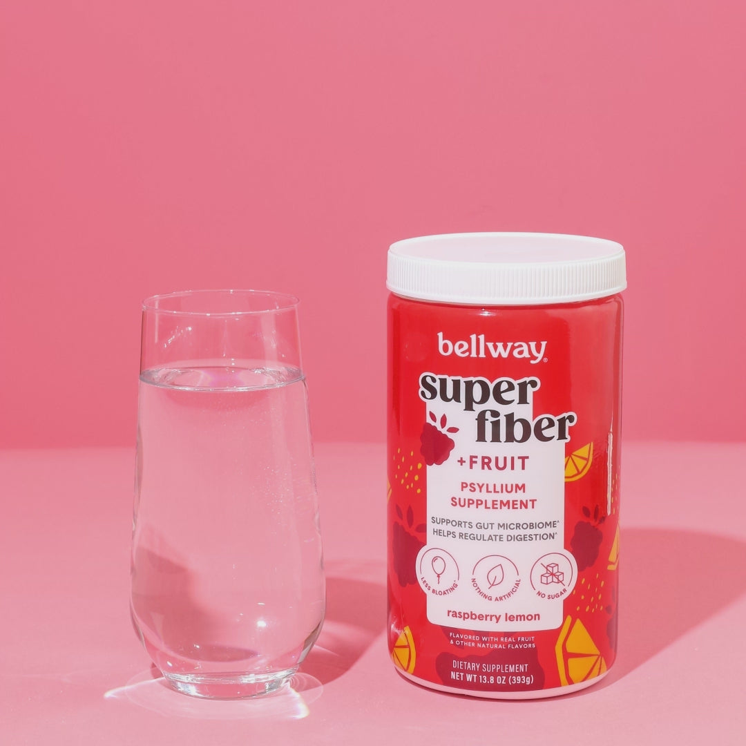 Bellway Super Fiber + Fruit raspberry lemon tub is pictured with a clear glass of water. A hand comes in and scoops the fiber supplement from the tub, puts it in the glass of water, uses an electric frother to mix the drink, and takes the glass off screen. Text reads "just add water, froth, and enjoy!"